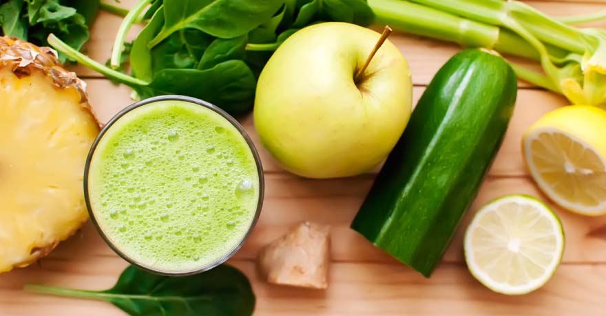 Green juice in cup, with yellow apple, cucumber and pineapple and lemon out on table